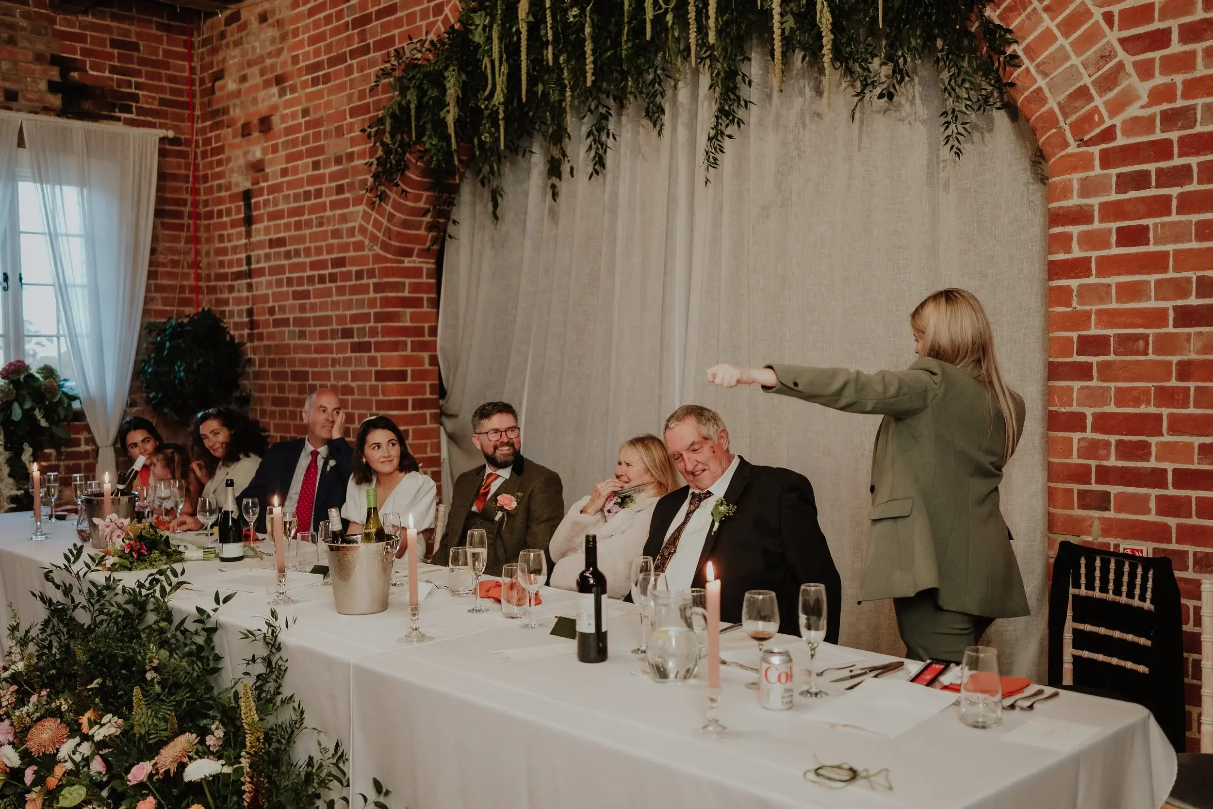 Top table guests laughing during speeches under a garland of trailing amaranthus in the exposed brick barn
