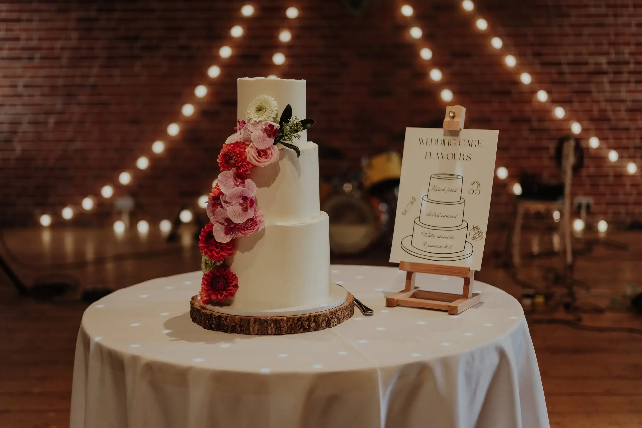 Three tiered white wedding cake trimmed with pink orchids and dahlias, beside a hand-lettered flavours sign strung with festoon lights