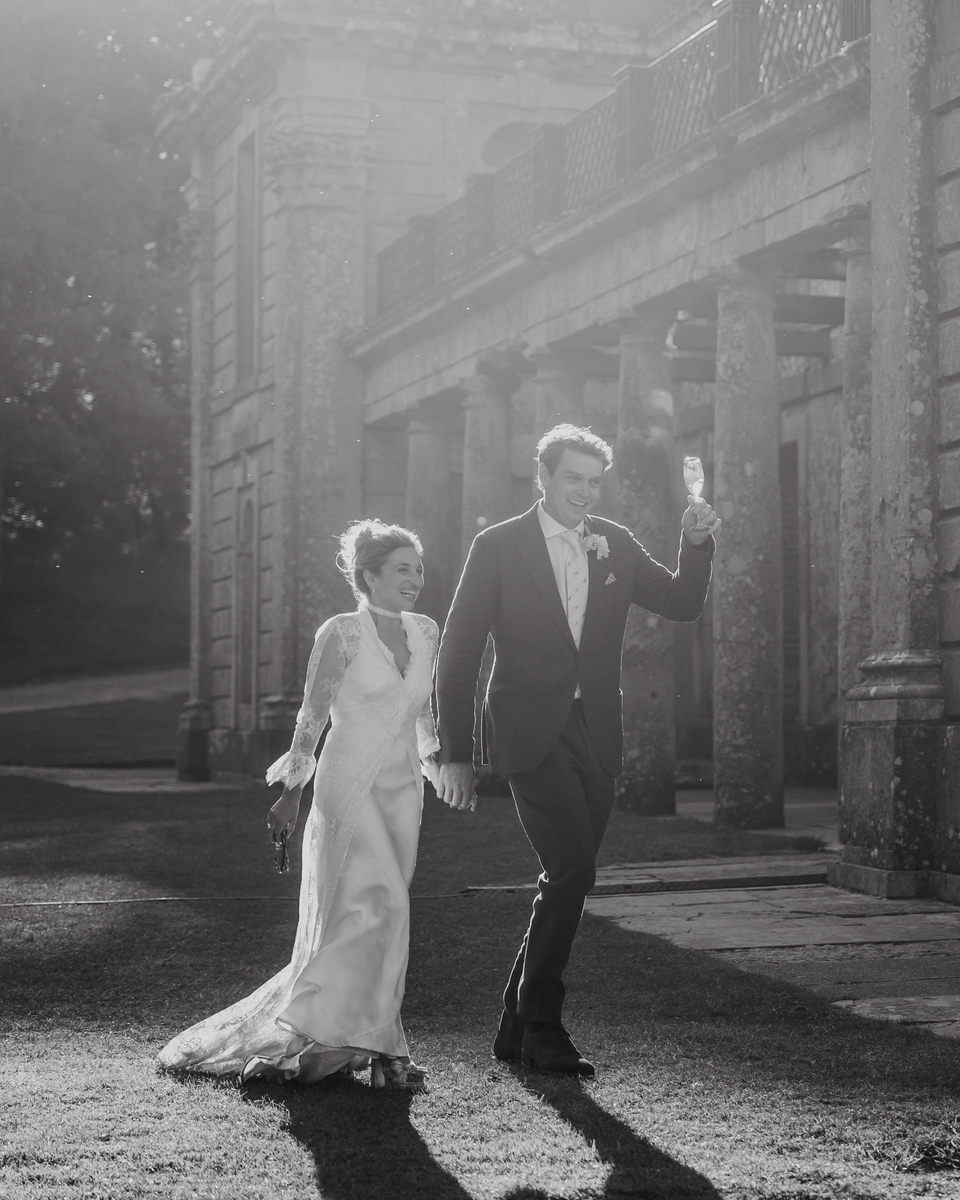 Bride and groom walking past the stone portico of Appuldurcombe House with a champagne toast