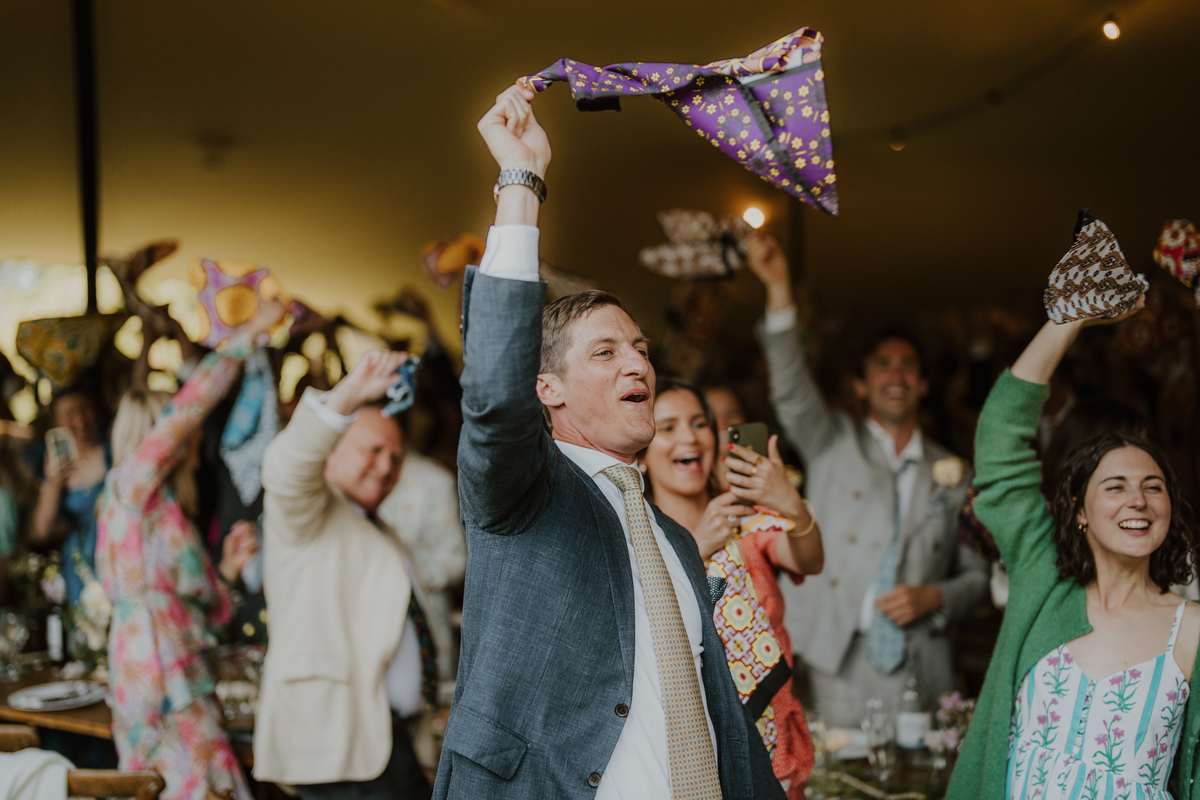Groom standing and waving a patterned napkin above his head as guests cheer behind him