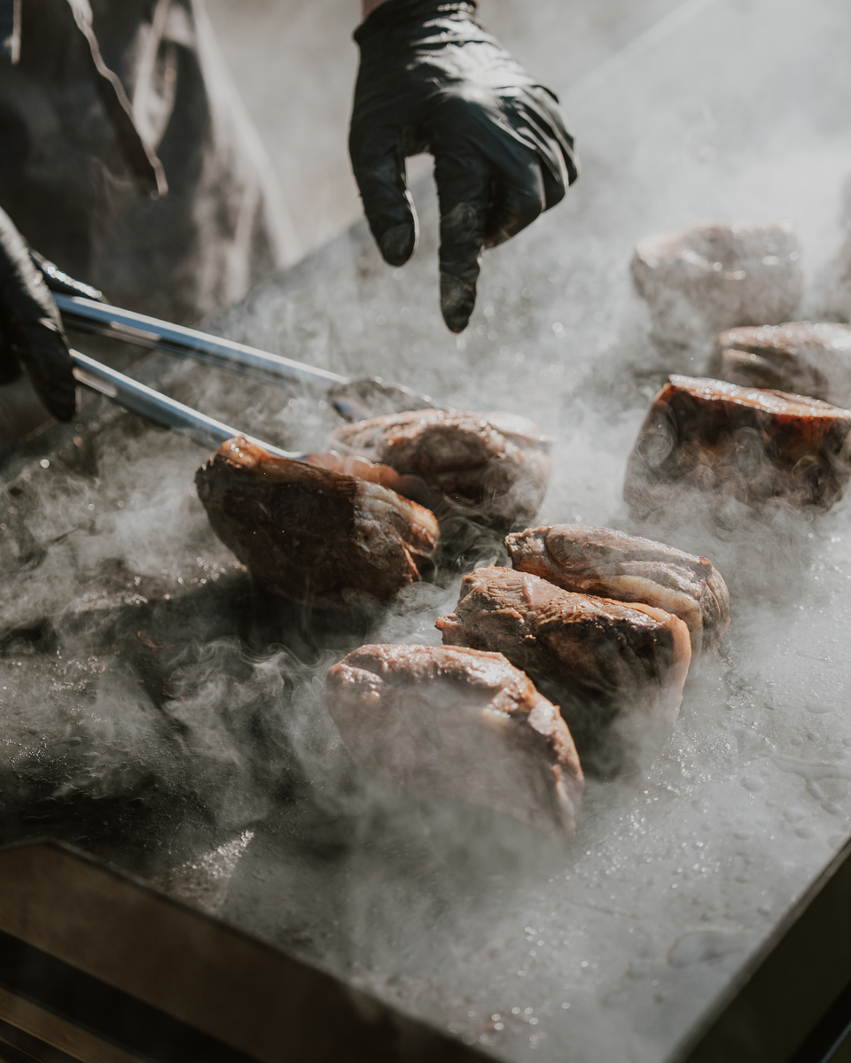 Chef searing steaks on a hot plate with billowing smoke and black gloved hands