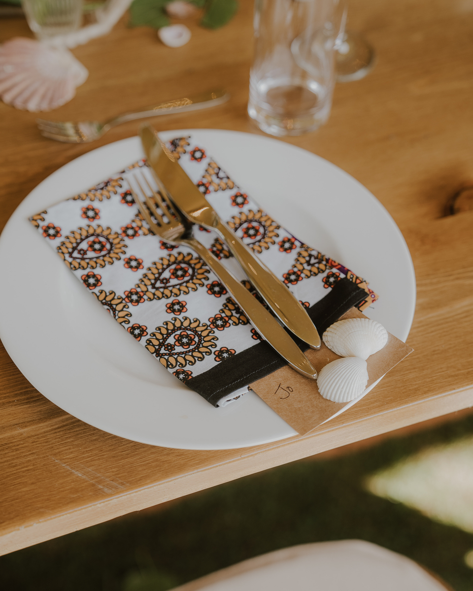 Place setting with gold cutlery on a paisley napkin tied with a name card and seashells