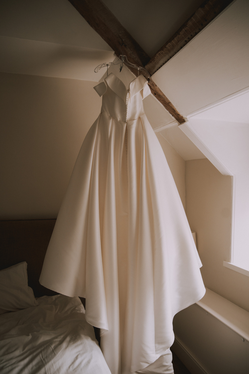 Off-the-shoulder satin wedding gown hanging from a wooden beam in an attic bedroom, its full skirt pooling over the bed below