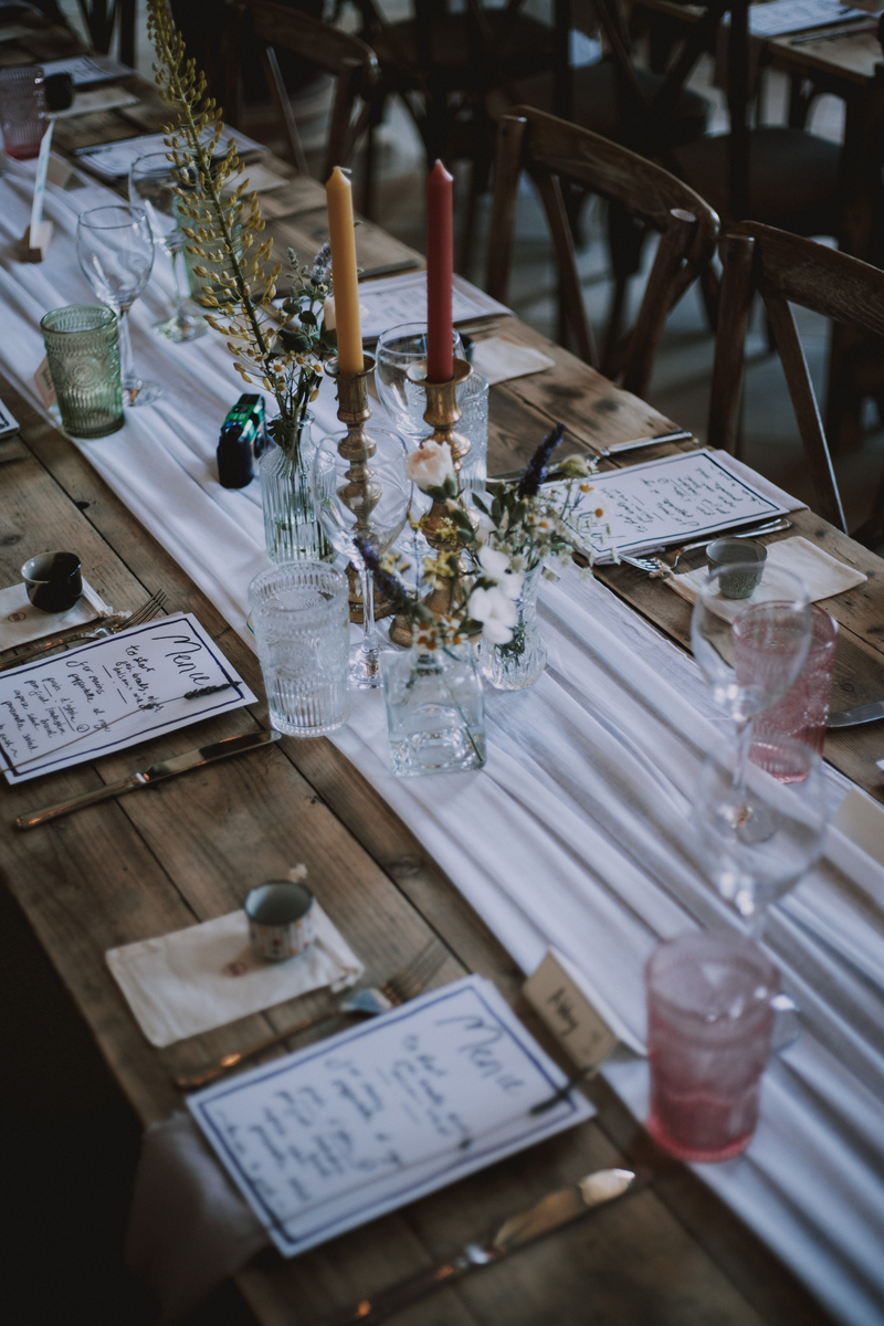 Long feasting table set with mismatched glassware, brass candlesticks and wildflowers in glass bottles down a white runner