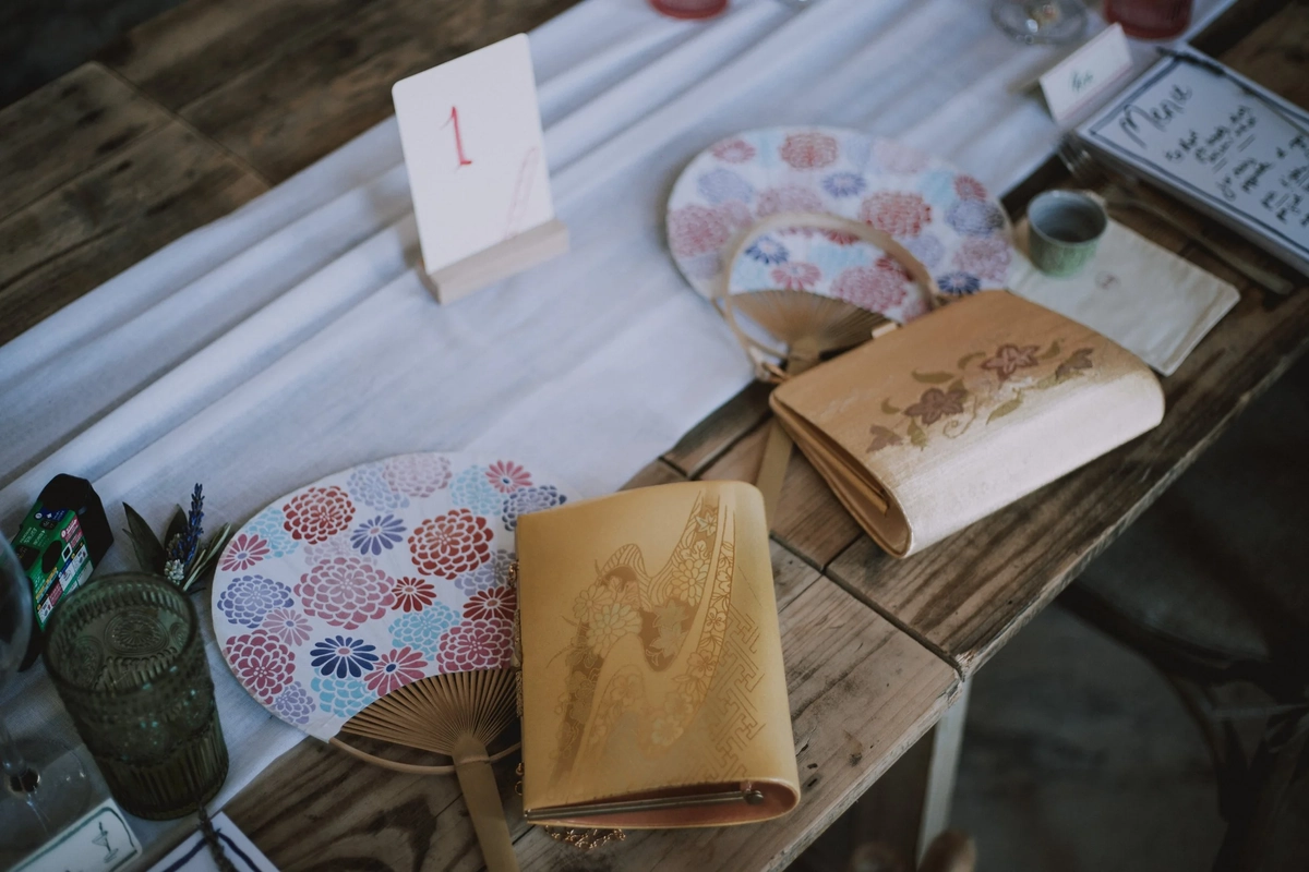 Painted fans and gilded clutch bags laid out on a rustic table beside a hand-lettered table number and menu card