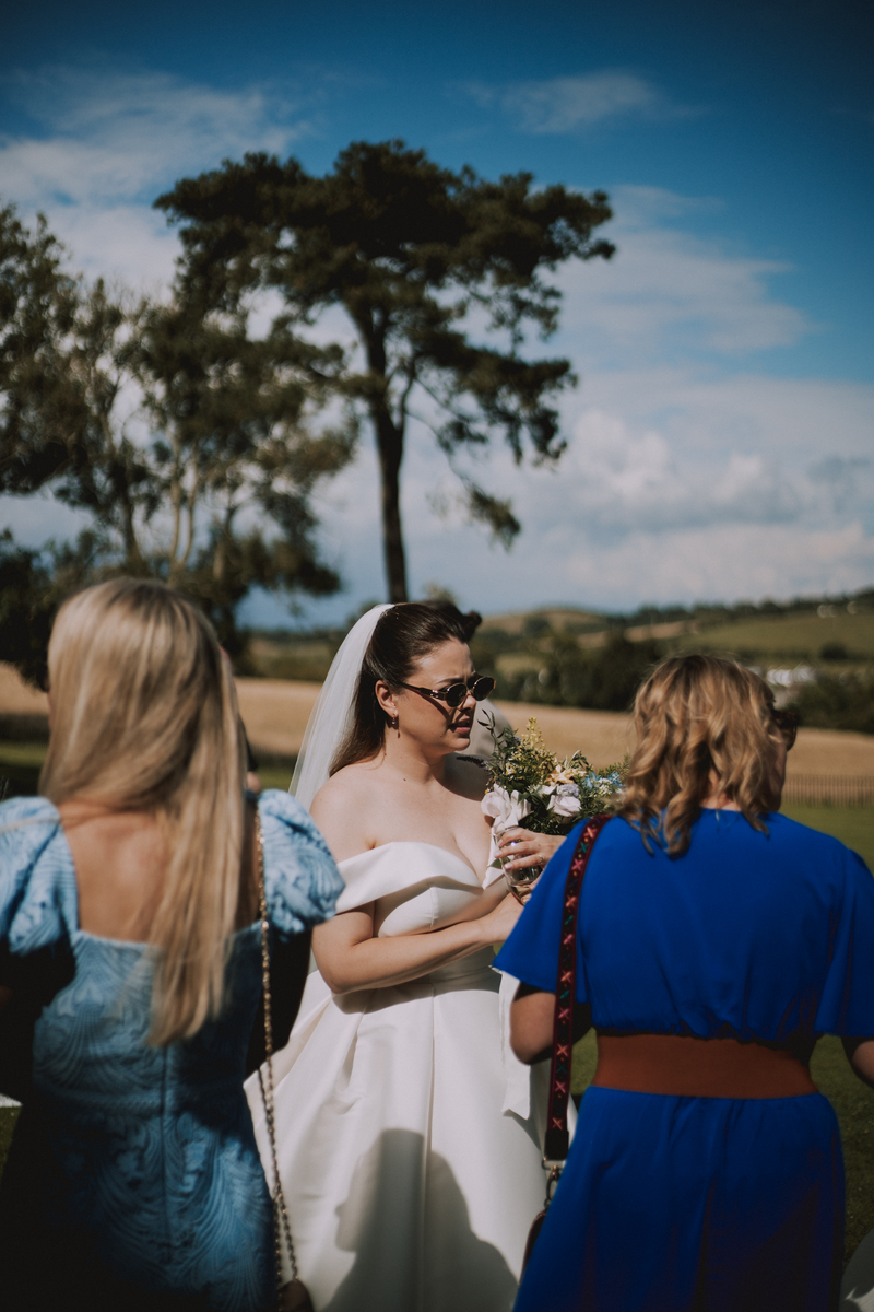 Rebecca in sunglasses and veil chats with bridesmaids in blue dresses on the lawn, rolling countryside behind them