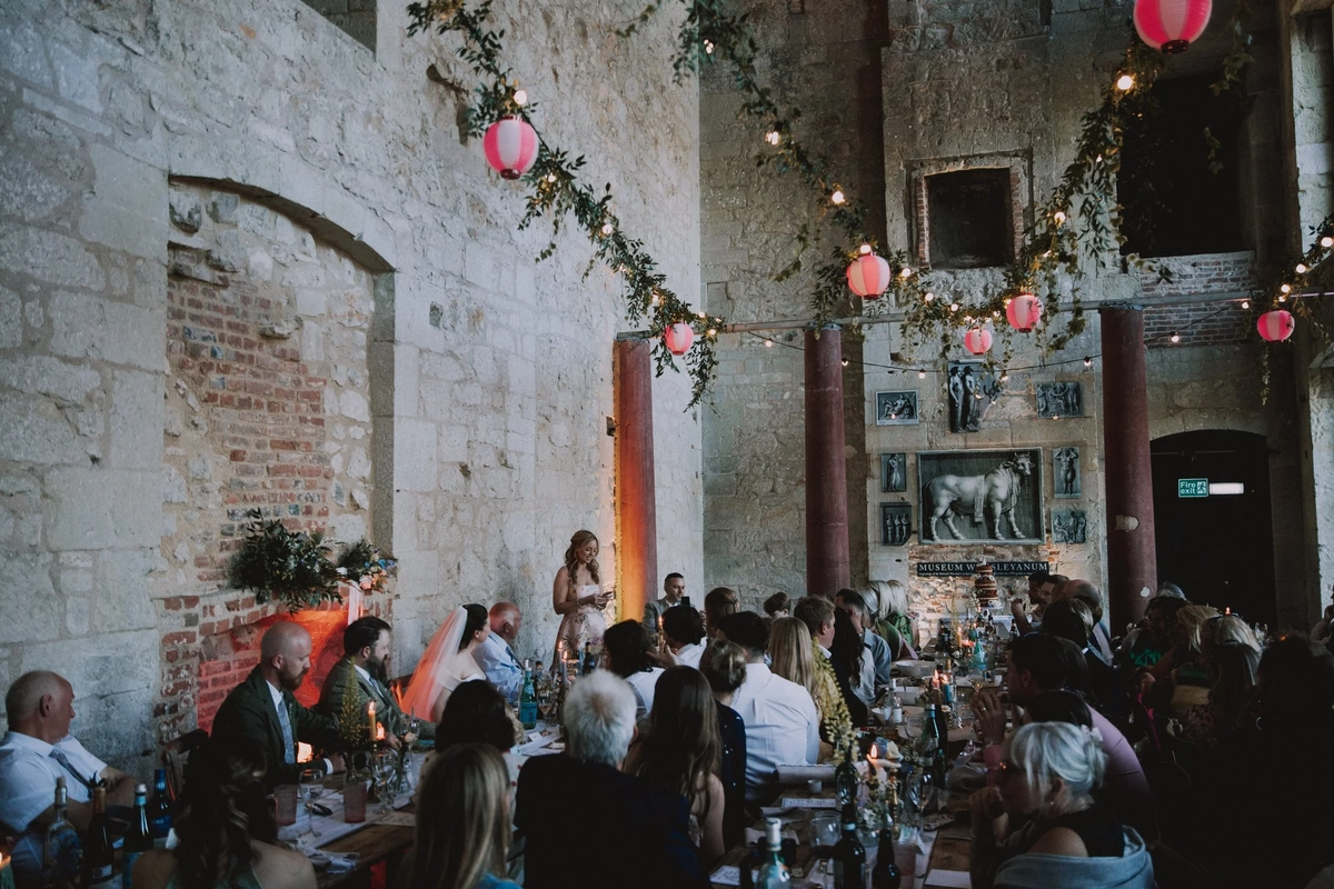A bridesmaid gives a speech beside the newlyweds' table, paper lanterns and fairy lights glowing overhead in the ruins