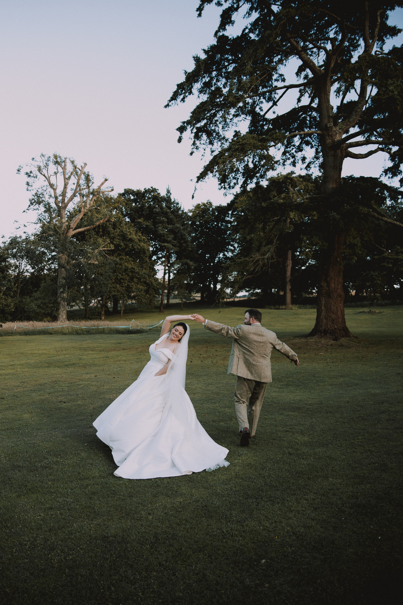 Russel spins Rebecca across the parkland lawn at dusk, her gown swirling, tall cedar trees silhouetted behind them
