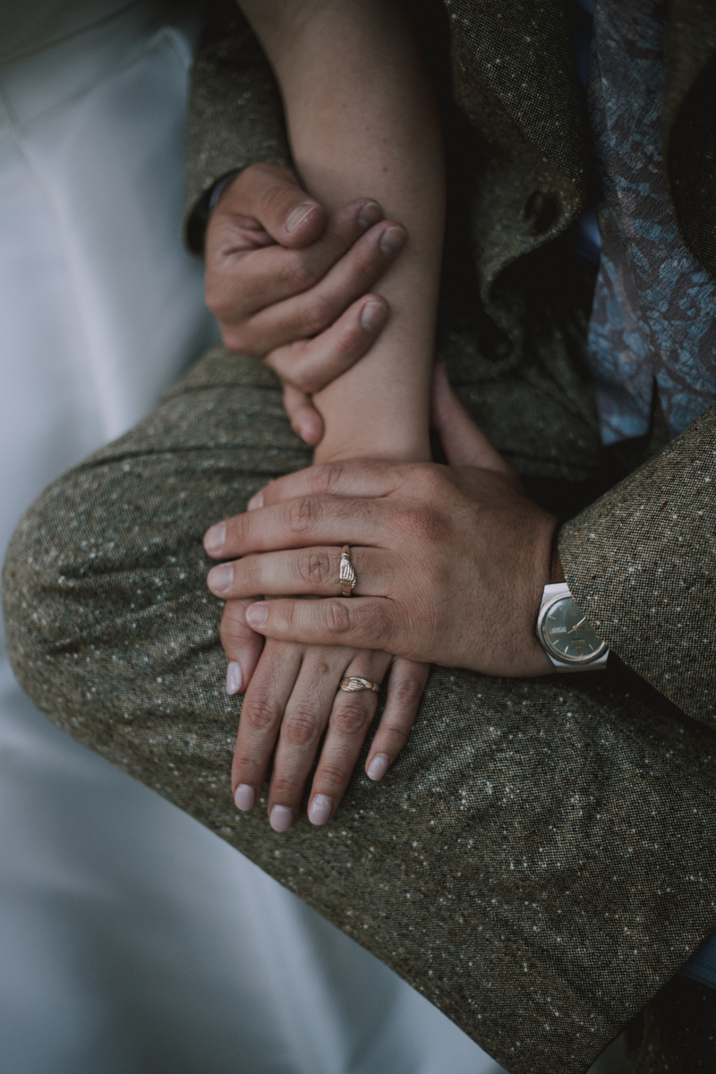 Close-up of the couple's clasped hands resting on tweed, new wedding rings and a watch catching the light