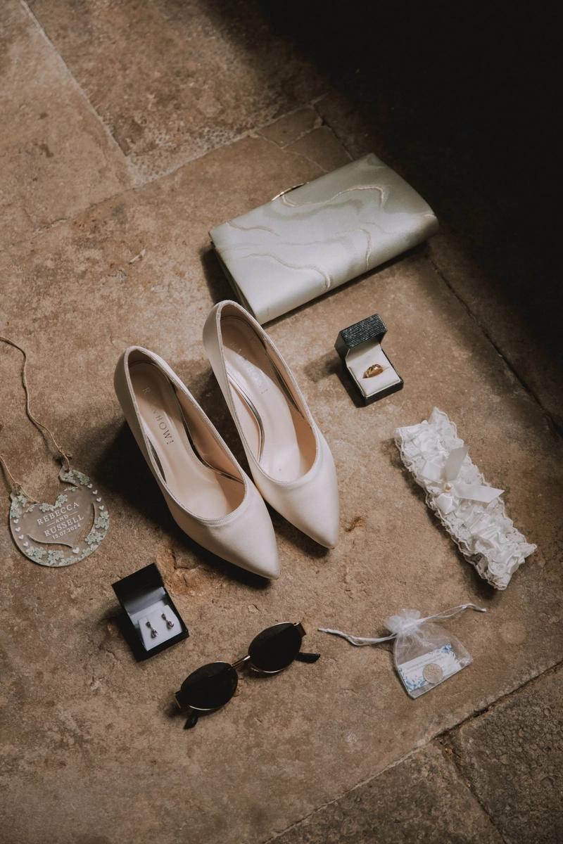 Bridal details laid on stone: satin heels, a marbled clutch, a garland pin ring box, earrings, sunglasses and a heart-shaped keepsake