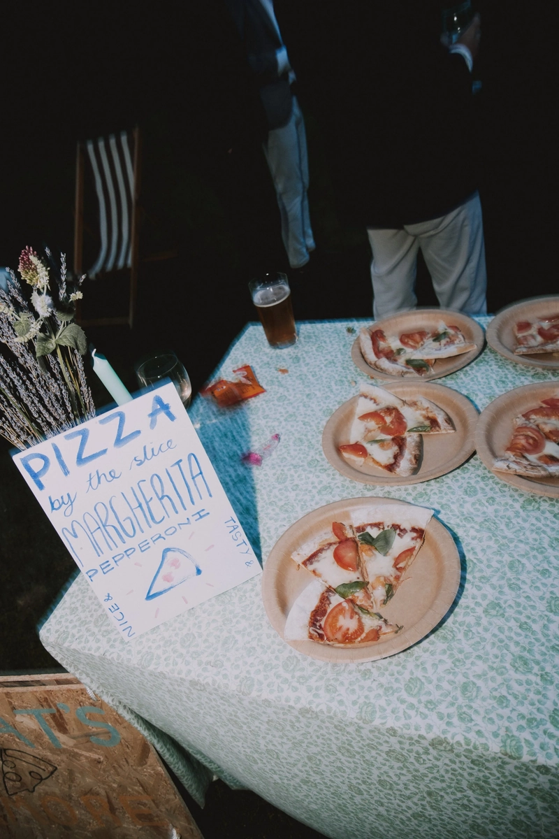 Handwritten sign for pizza by the slice beside paper plates of margherita and pepperoni on a floral-cloth table at dusk