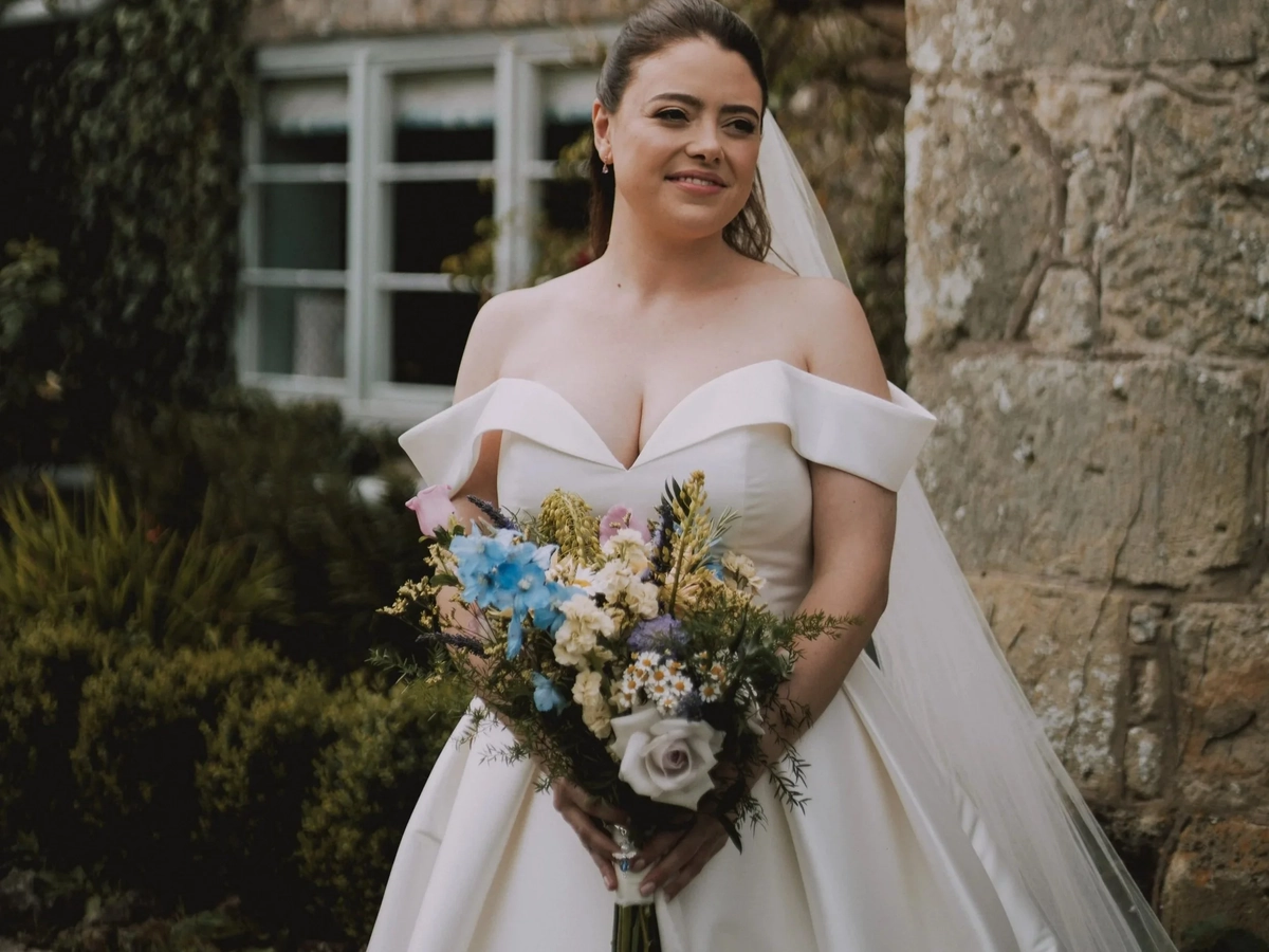 Rebecca in her off-the-shoulder gown and veil holding a wild bouquet of blue delphiniums and lilac roses against an ivy-covered stone wall