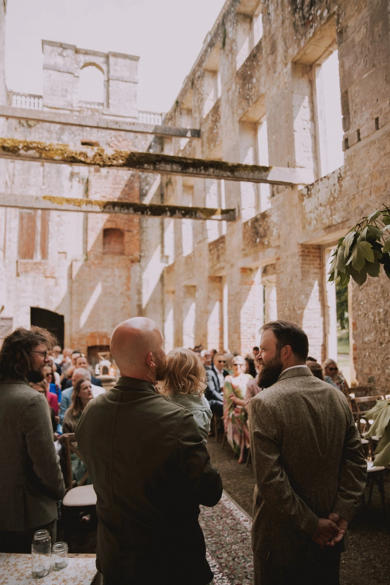 Guests in tweed and linen stand facing the open-air ceremony inside the roofless stone ruins of Appuldurcombe House on the Isle of Wight