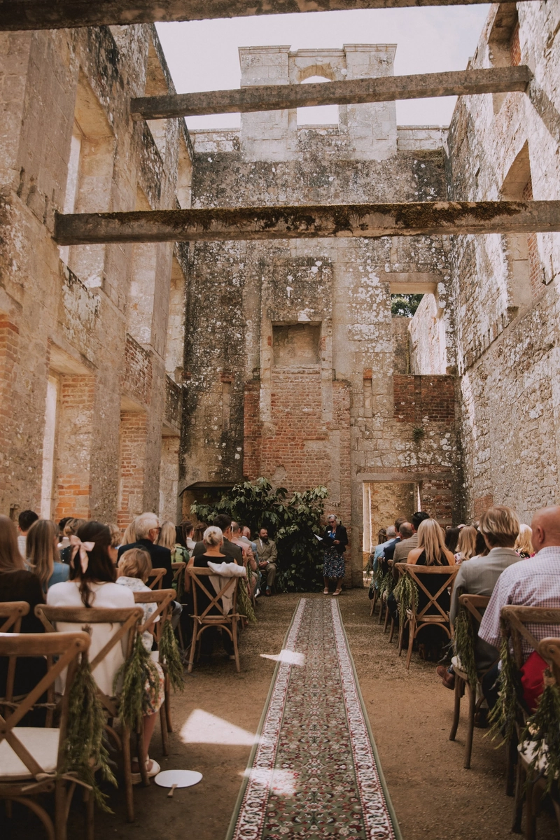 Rows of chairs dressed with greenery lead up a patterned runner between the towering weathered walls of the roofless ceremony ruins