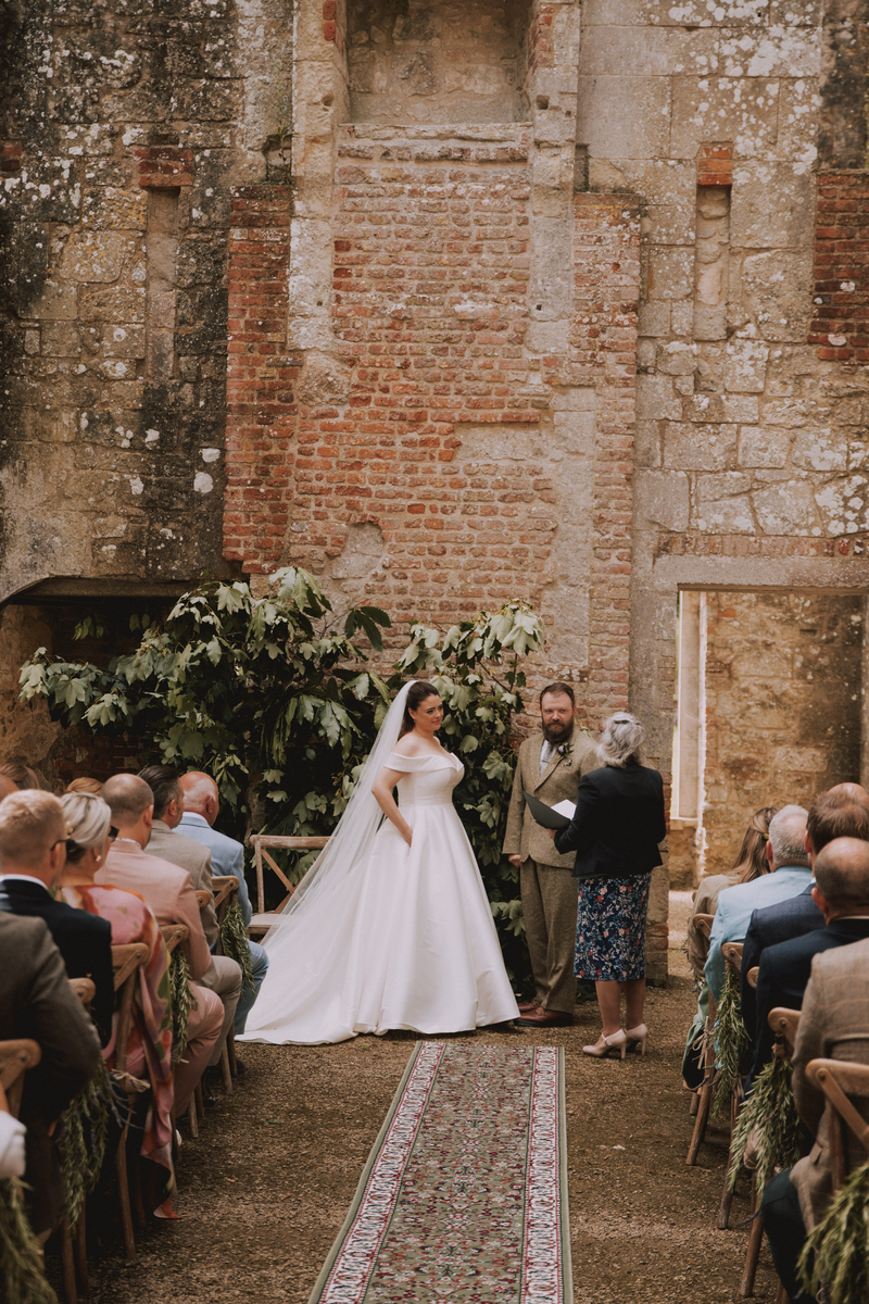 Rebecca in her ballgown stands at the altar with Russel and the celebrant, framed by potted trees against exposed brick and stone