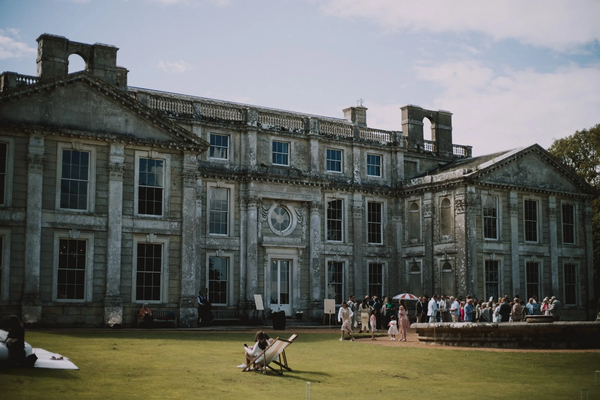 Wide view of the baroque facade of Appuldurcombe House with guests gathered on the lawn and two deckchairs set out front