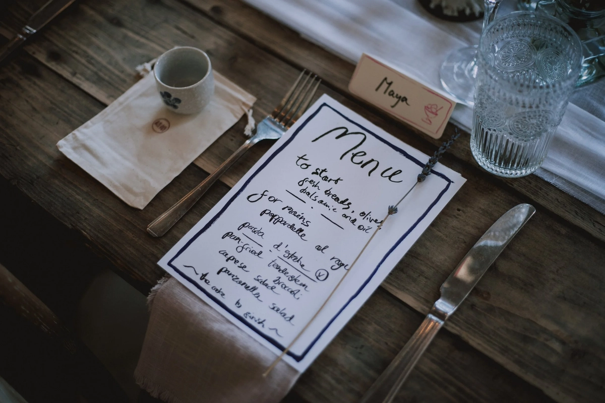 Handwritten menu card promising pappardelle al ragu and panzanella salad set beside a linen napkin and cut-glass tumbler