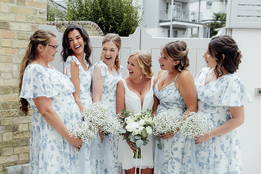 Rosie's bridesmaids in matching blue floral dresses gathered round her, all laughing together and holding gyp bouquets