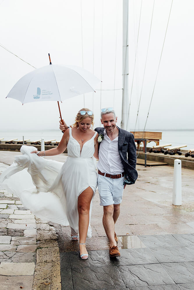 Rosie and Adam walking together under a white umbrella past mooring posts on the Isle of Wight waterfront, her dress caught in the breeze