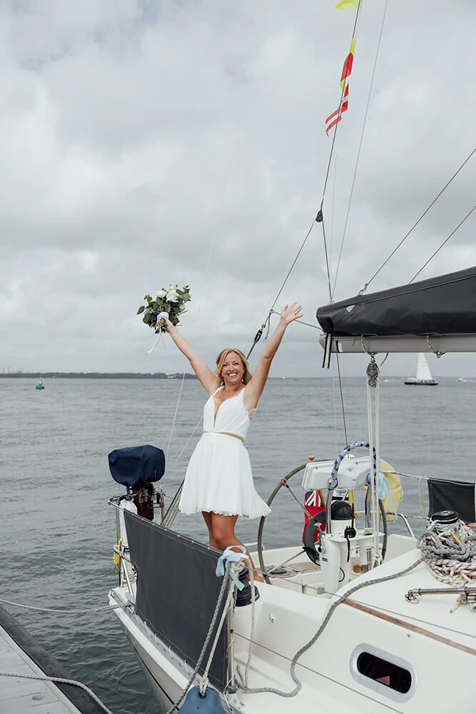 Rosie punching the air with her bouquet raised on the bow of the sailing yacht, a second yacht passing on the water beyond