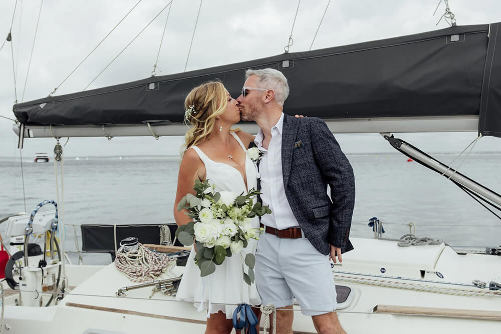 Rosie and Adam kissing on the yacht deck beneath the boom, her bouquet of white roses and eucalyptus between them