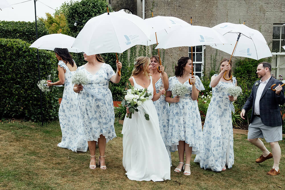 Rosie and her bridesmaids walking together under white umbrellas in the garden, all in matching blue floral dresses