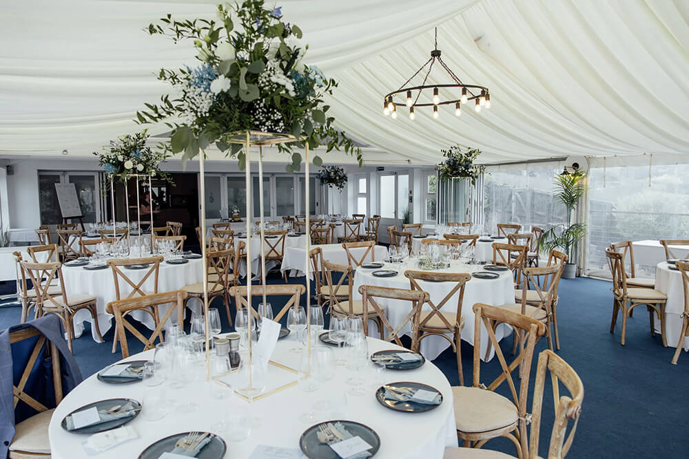 Marquee reception room set with round tables, crossback chairs and tall floral arrangements beneath a wagon-wheel chandelier