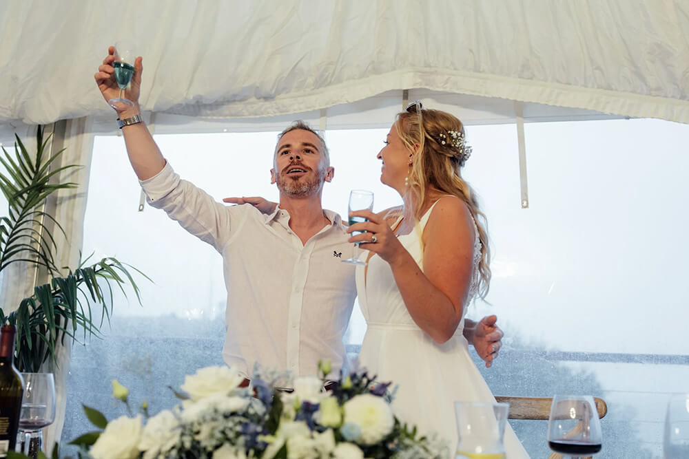 Adam raising a glass to give a speech under the marquee awning as Rosie smiles beside him with her own drink in hand