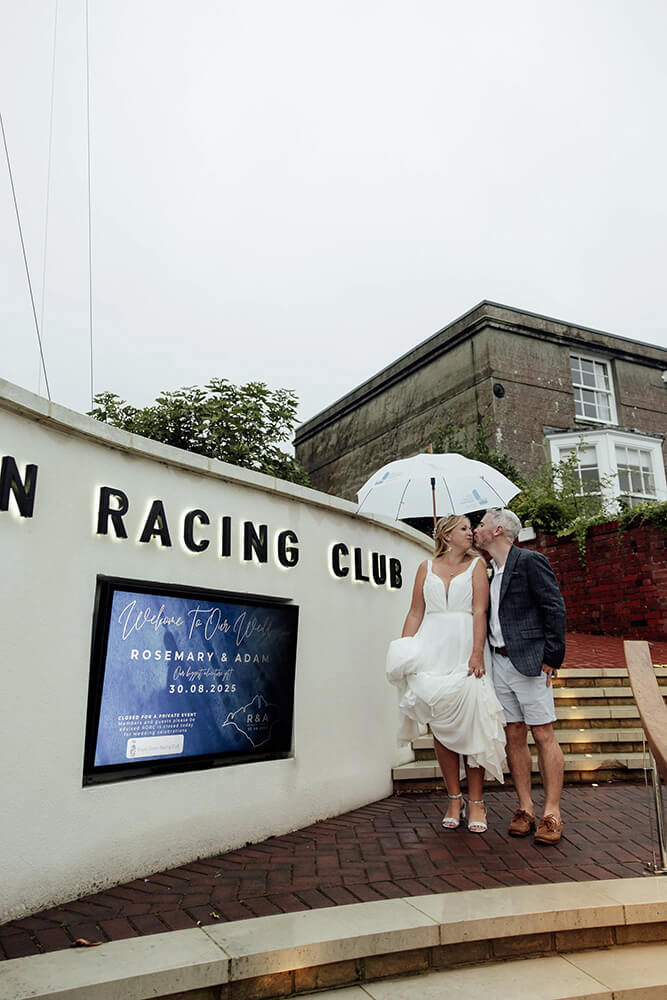 Rosie and Adam sharing a kiss under an umbrella beside the illuminated Royal Ocean Racing Club sign welcoming their wedding guests