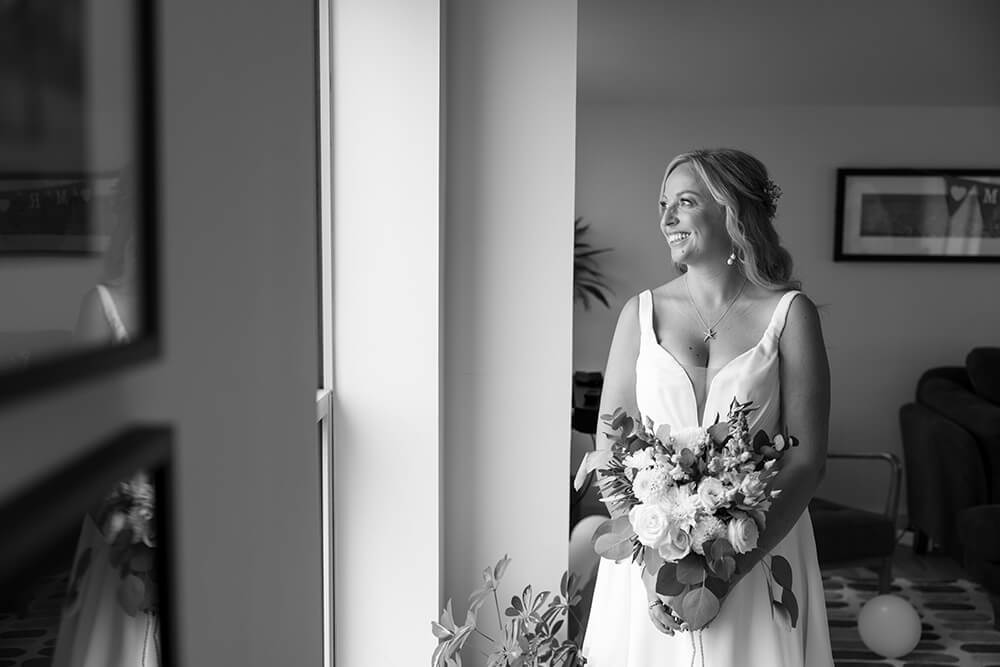 Rosie glancing out of a window in soft daylight, her bouquet of white roses and dahlias held at her waist before the ceremony