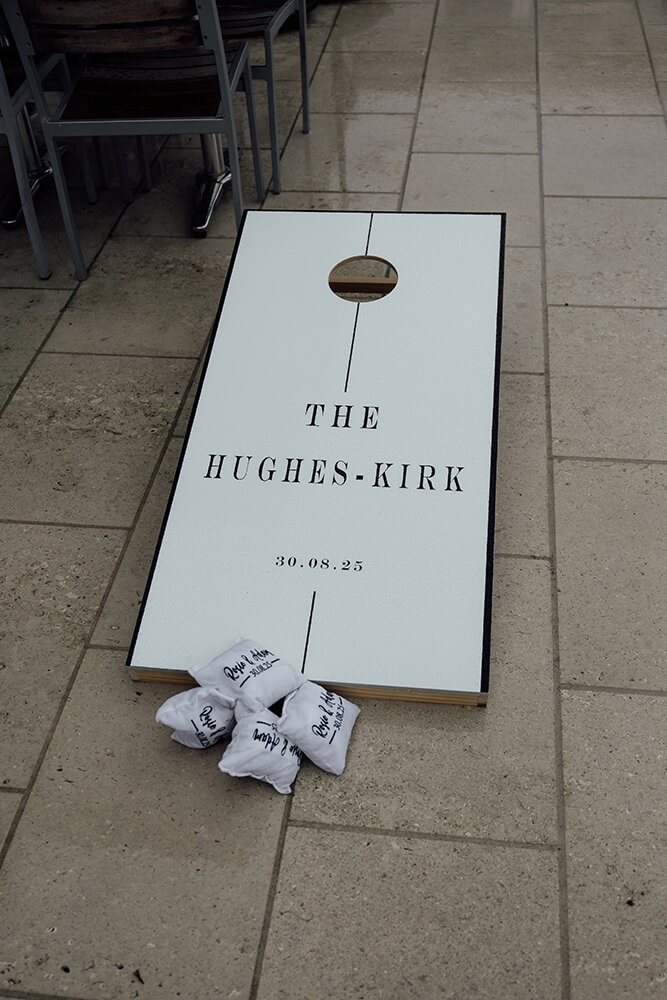 A personalised cornhole board reading "The Hughes-Kirk 30.08.25" set out on the terrace with monogrammed bean bags