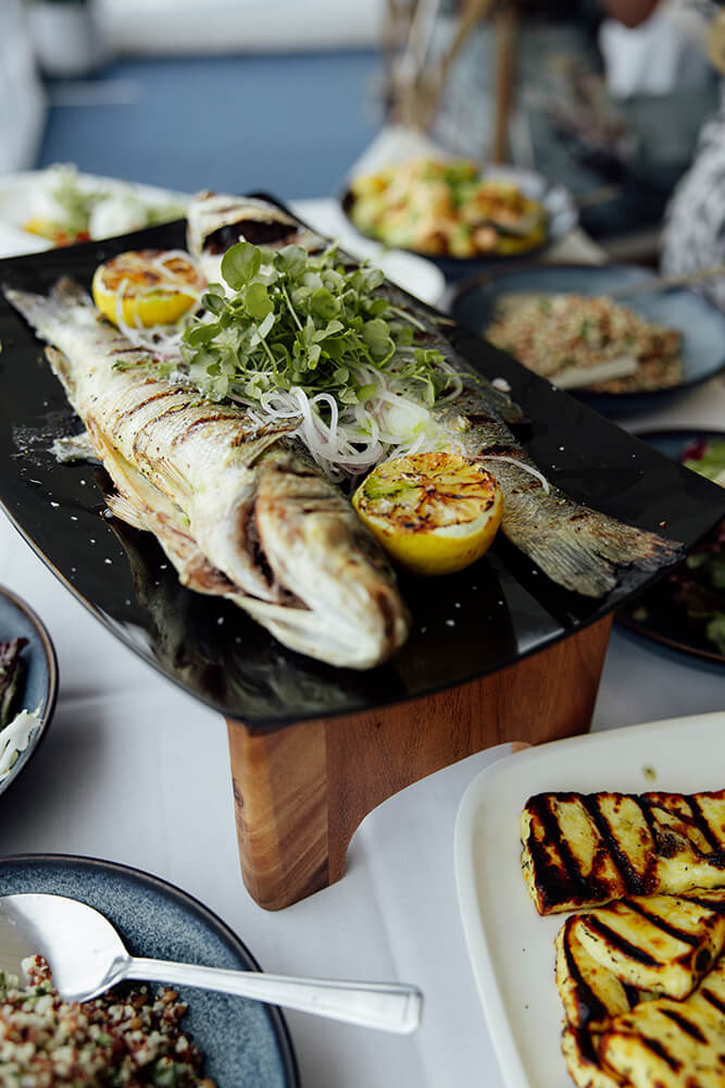 Whole grilled sea bream with charred lemon halves and watercress served on a wooden board alongside grilled halloumi