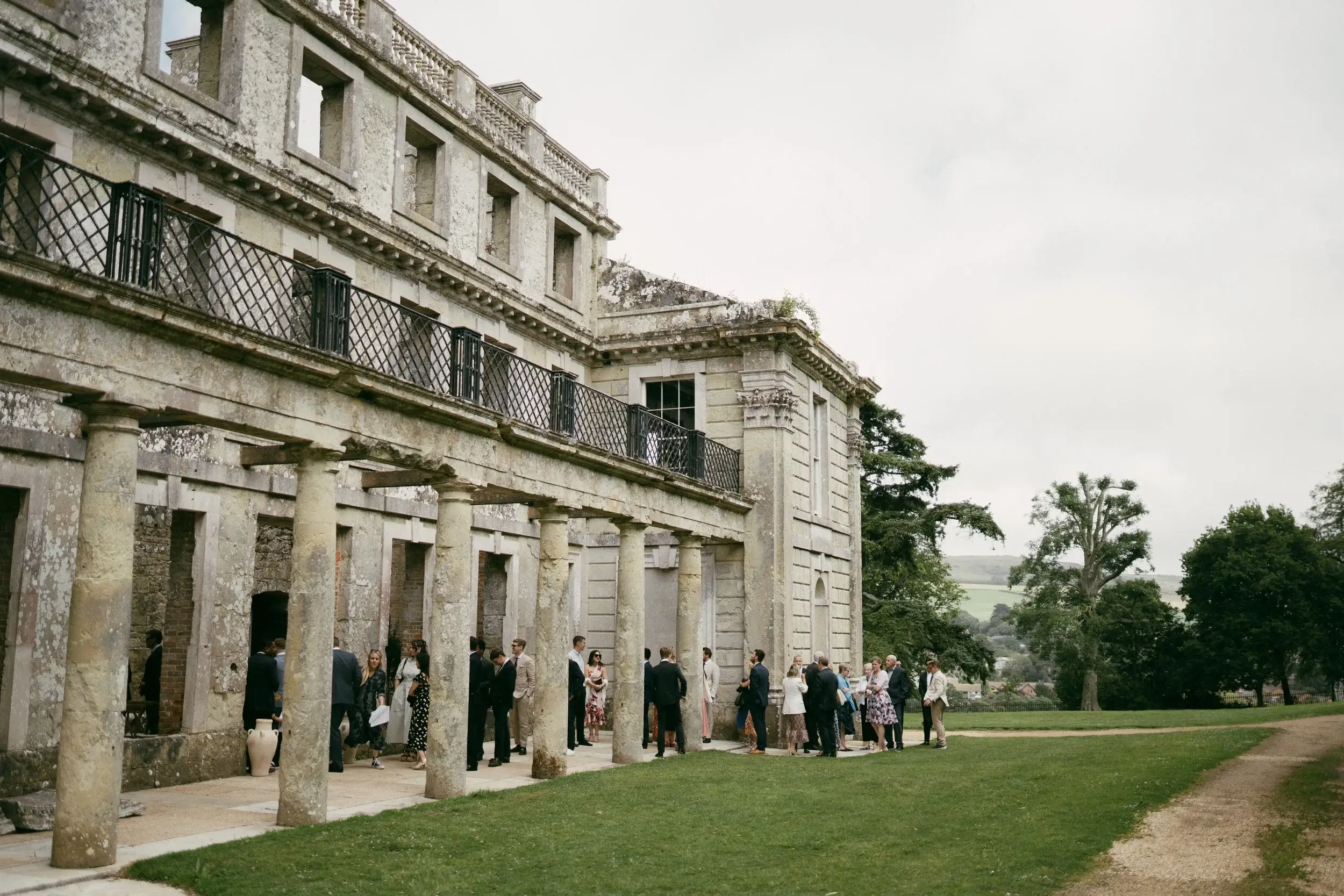 Guests gathering under the colonnade of Appuldurcombe House, the baroque ruin's stone columns framing the Isle of Wight countryside beyond