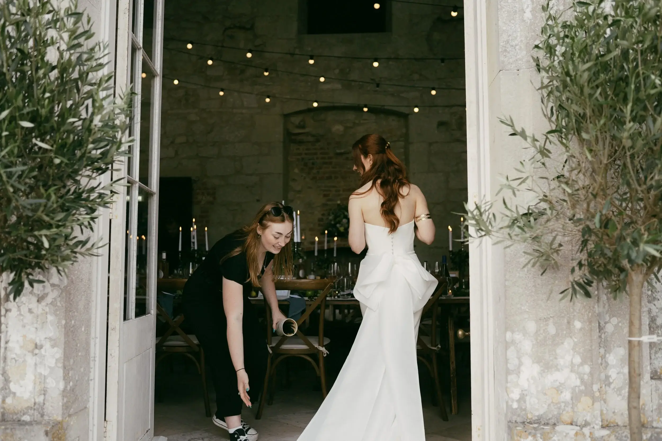 A stylist adjusts the bow on the back of Rosie's wedding dress as she stands in the Great Hall doorway framed by olive trees