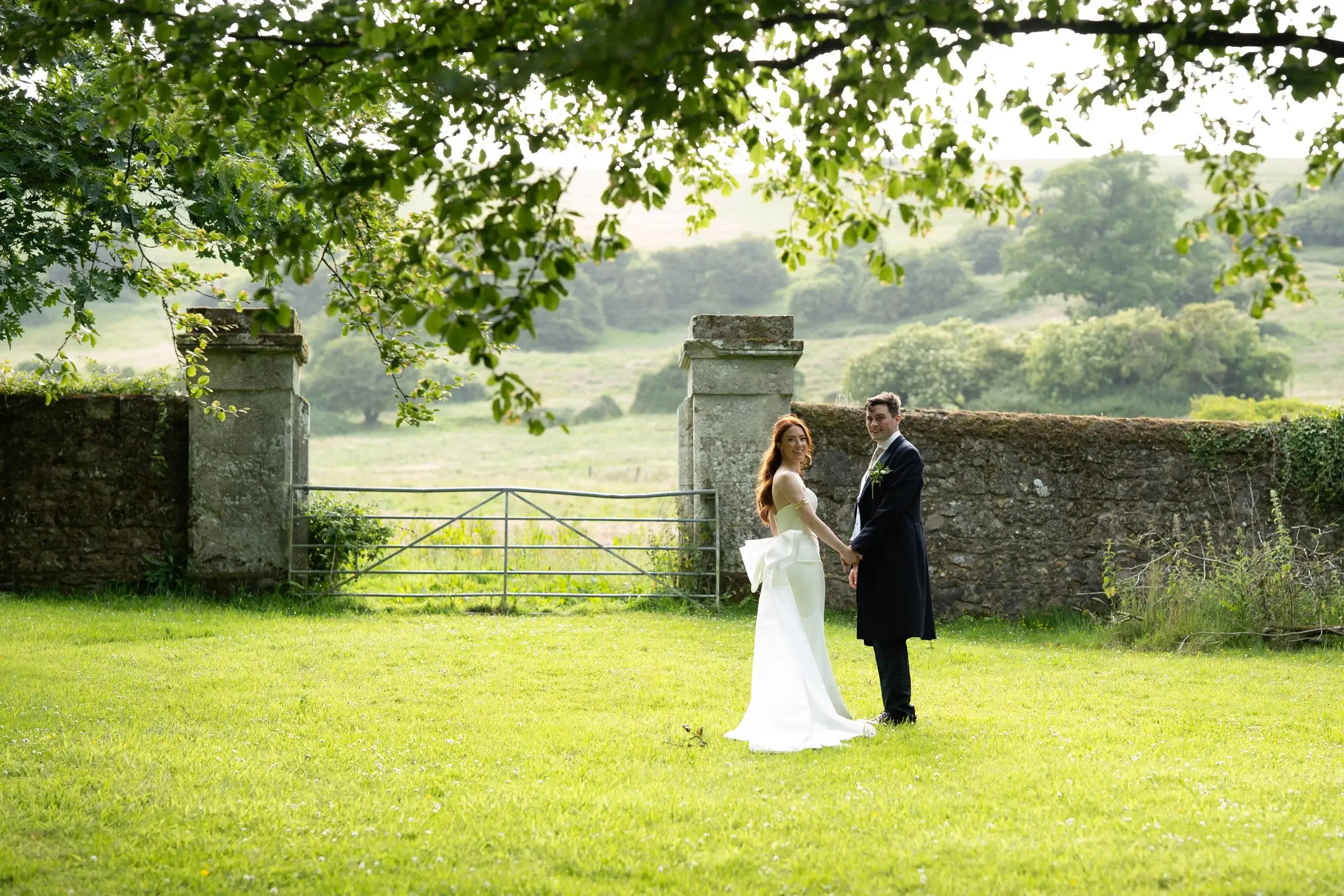 Rosie and George hold hands on the lawn beneath a tree, an old stone gateway and rolling Isle of Wight hills behind them