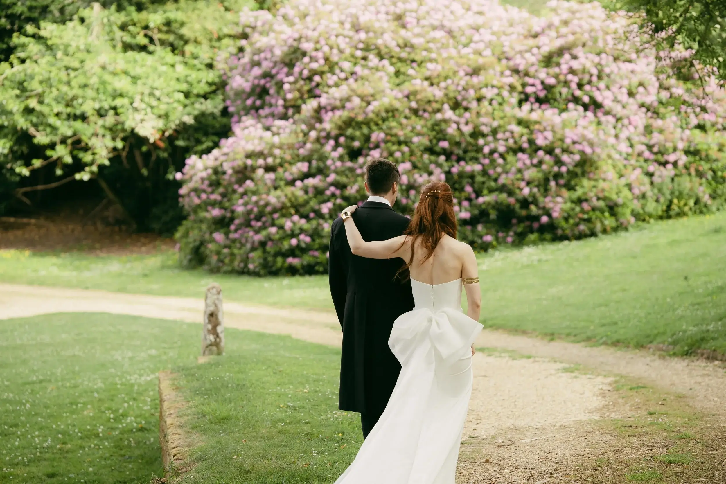 Rosie rests her hand on George's shoulder as they walk together past a blooming pink rhododendron hedge