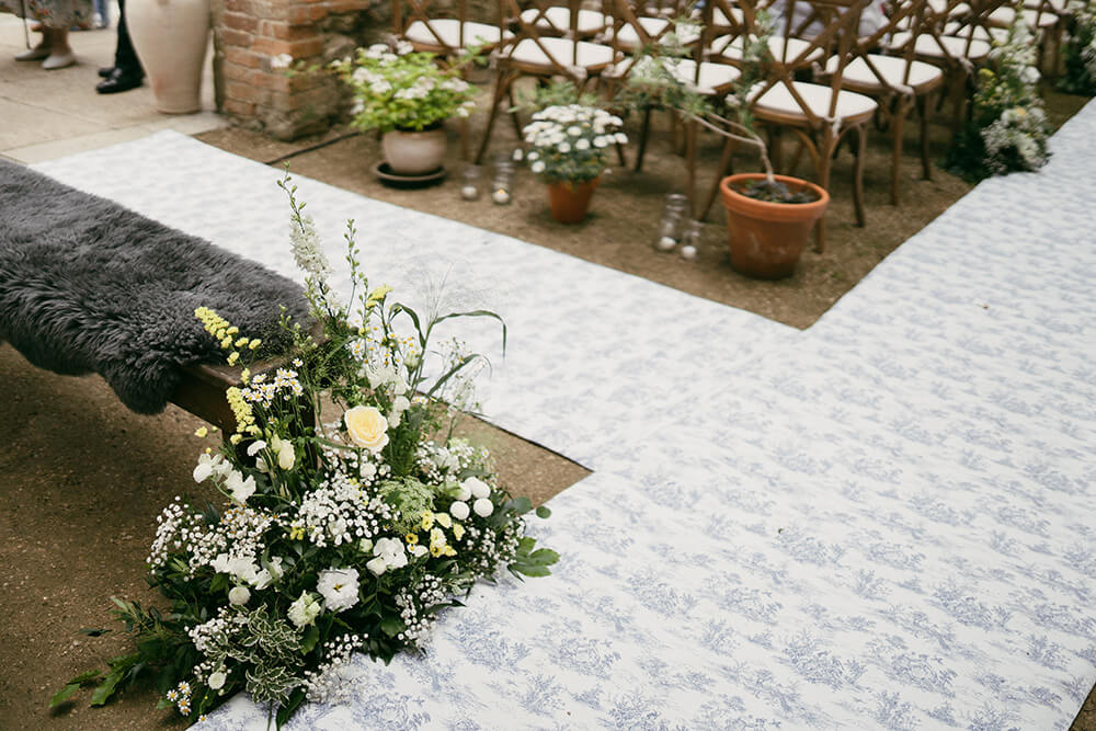 Bridal bouquet of white roses and wildflowers laid on a sheepskin-draped bench beside the aisle runner and potted florals