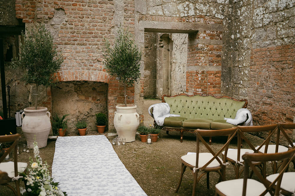 Vintage green velvet sofa and olive trees in urns set up beside the aisle in the roofless ruins ahead of the ceremony