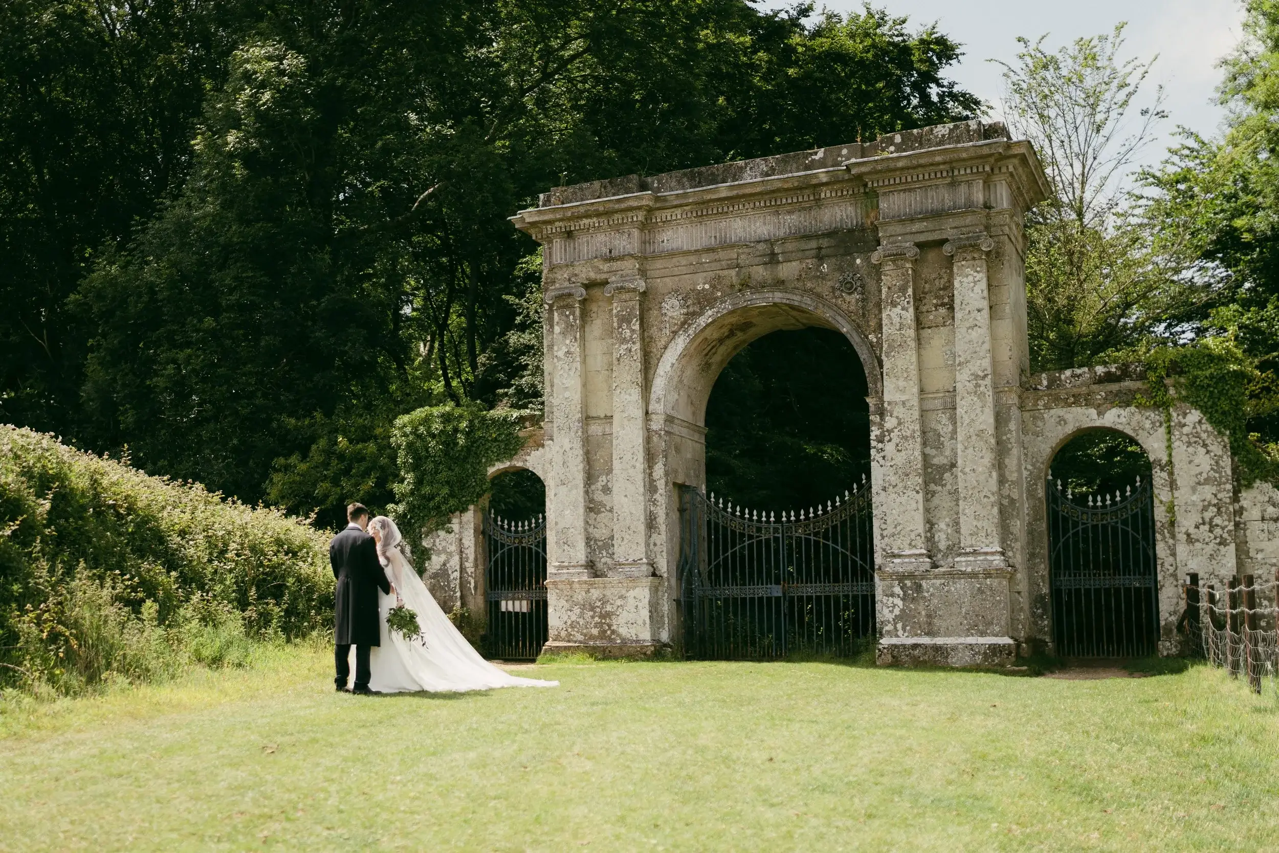 Rosie and George stand arm in arm before the towering stone archway and ironwork gates on the Appuldurcombe House grounds