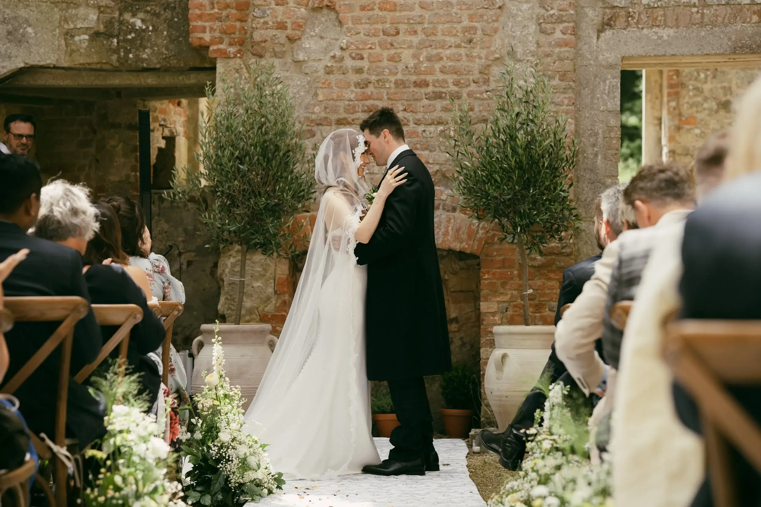 Rosie and George share a first embrace at the altar between two potted olive trees against a weathered brick wall