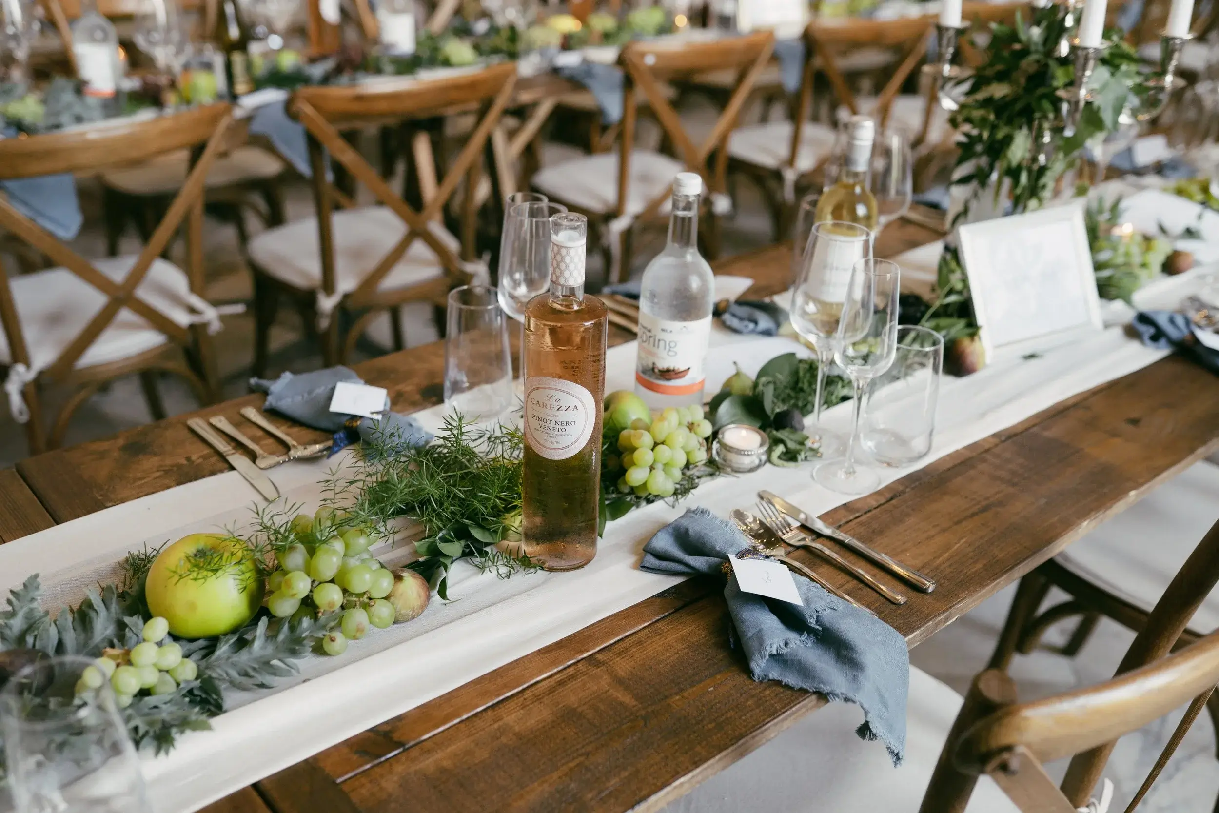 Close-up of a feasting table with rosé and sparkling water bottles, grapes, apples and rosemary sprigs on a linen runner