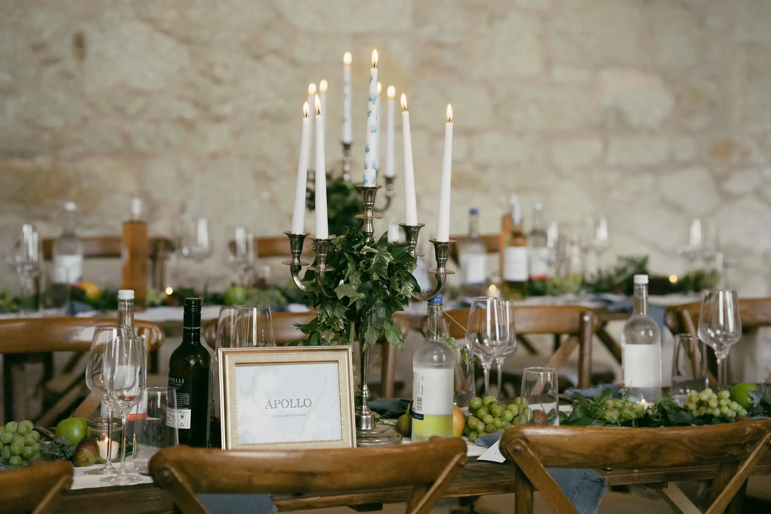 Silver candelabra wrapped in ivy beside a table sign reading Apollo, with wine bottles and grapes on the feasting table